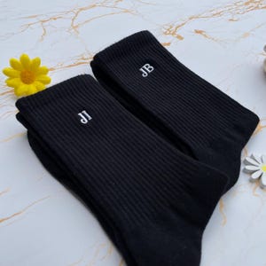 May include: A pair of black ribbed socks with white embroidered initials. One sock displays "JI" and the other "JB". A yellow flower and a white daisy-shaped decoration are in the background.