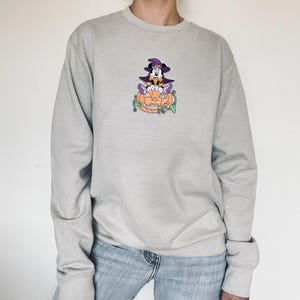 Pu&ograve; includere: Felpa grigio chiaro con un design ricamato a tema Halloween. Il design include un personaggio dei cartoni animati con un cappello da strega, seduto su una zucca intagliata con viti verdi. La felpa ha maniche lunghe e girocollo.