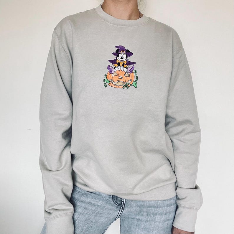 Pu&ograve; includere: Felpa grigio chiaro con un design ricamato a tema Halloween. Il design include un personaggio dei cartoni animati con un cappello da strega, seduto su una zucca intagliata con viti verdi. La felpa ha maniche lunghe e girocollo.