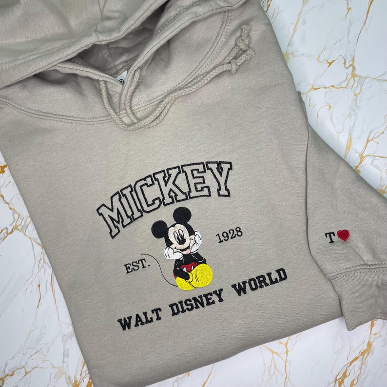 May include: A beige hoodie featuring the word "MICKEY" in black lettering, along with an image of Mickey Mouse and the text "EST. 1928 WALT DISNEY WORLD." The sleeve has the letter "T" and a red heart. The hoodie has a drawstring hood.