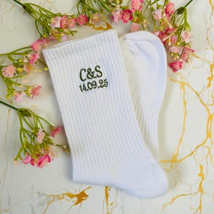 May include: Two white ribbed socks, embroidered with the initials "C&S" and the date "14.09.25" in green. The socks are presented on a surface with a marble effect, with pink and white flowers arranged around them.