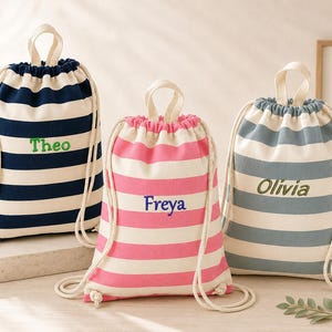 May include: Three drawstring backpacks made of canvas fabric with horizontal stripes. The left bag is navy blue and white with the name Theo embroidered in green. The center bag is pink and white with the name Freya in blue. The right bag is grey and white with the name Olivia in green.
