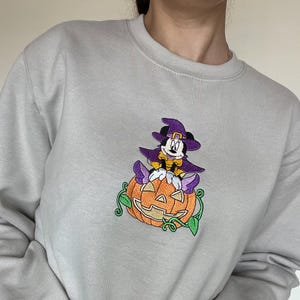 Pu&ograve; includere: Felpa grigio chiaro con un ricamo a tema Halloween. Il design presenta un personaggio dei cartoni animati con un cappello e un mantello da strega viola, seduto su una zucca arancione con viti verdi.