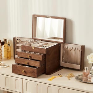 May include: A wooden jewelry box with multiple drawers and compartments. The box is open, revealing a mirror, drawers, and storage for necklaces and rings. The box is made of dark wood and has gold-colored hardware. The jewelry box is on a white surface.