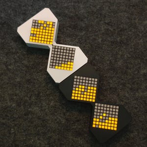 Può includere: Quattro pannelli luminosi a LED modulari geometrici in fila. I pannelli sono bianchi e neri, con una griglia di piccoli quadrati che si illuminano in giallo e grigio. I pannelli sono disposti su una superficie grigio scuro.
