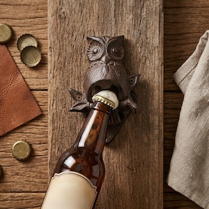 May include: A cast iron owl bottle opener mounted on a wooden plank. The owl is dark brown with detailed features, holding a bottle cap in its beak. A brown beer bottle is being opened. The background includes a wooden surface, bottle caps, and a tan cloth.