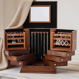Puede incluir: Un joyero de madera oscura con múltiples cajones extraíbles, compartimentos laterales para pendientes y un soporte central para collares. El interior cuenta con un forro de terciopelo negro, un espejo rectangular y pomos de metal dorado.