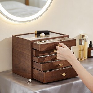 May include: A dark wood jewelry box with multiple drawers and a glass top. The drawers are being opened to reveal jewelry inside. The box has gold-colored knobs and sits on a reflective surface. A round mirror is in the background.