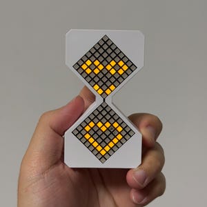 Könnte beinhalten: Eine weiße digitale Sanduhr-Uhr mit einer Gitteranzeige, die ein gelbes Herz zeigt. Die Uhr wird in einer Hand vor einem grauen Hintergrund gehalten. Die Anzeige besteht aus kleinen, leuchtenden Quadraten.