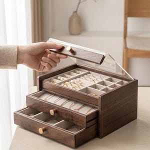 May include: A dark wood jewelry box with a clear acrylic lid and two pull-out drawers. The interior is lined with beige fabric, featuring compartments for rings and necklaces. Each drawer and the lid are fitted with small, round, gold-toned knobs.