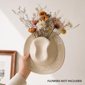 Op de afbeelding: Een crèmekleurige, aan de muur gemonteerde cowboyhoedvaas van keramiek, gevuld met gedroogde bloemen in oranje, geel, roze en wit. De hoed heeft een reliëfpatroon rond de rand. De tekst "FLOWERS NOT INCLUDED" is zichtbaar.