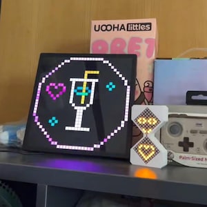 May include: A black square LED display with a pixelated cocktail graphic, a pink heart, and blue diamond shapes. A white hourglass-shaped LED display shows the numbers 12 and 2. A box of "UCOHA littles" is visible in the background.