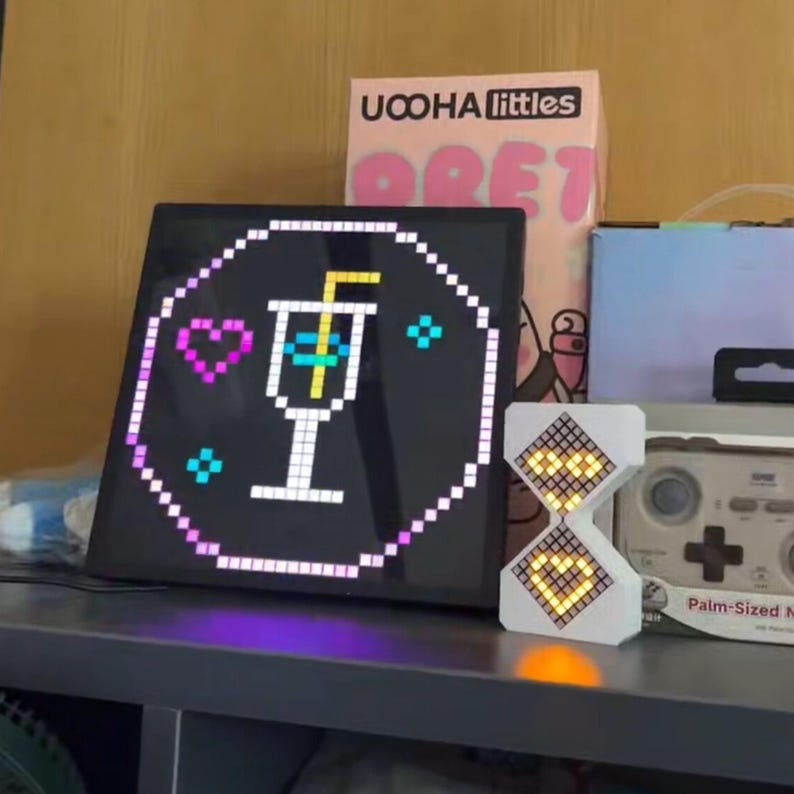May include: A black square LED display with a pixelated cocktail graphic, a pink heart, and blue diamond shapes. A white hourglass-shaped LED display shows the numbers 12 and 2. A box of "UCOHA littles" is visible in the background.