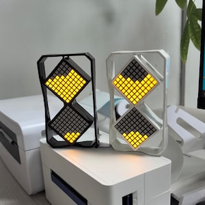 May include: Two digital clocks with a unique design. One clock has a black frame, the other is white. Each clock features two diamond-shaped displays with yellow and gray illuminated squares. The clocks are placed on a white surface.