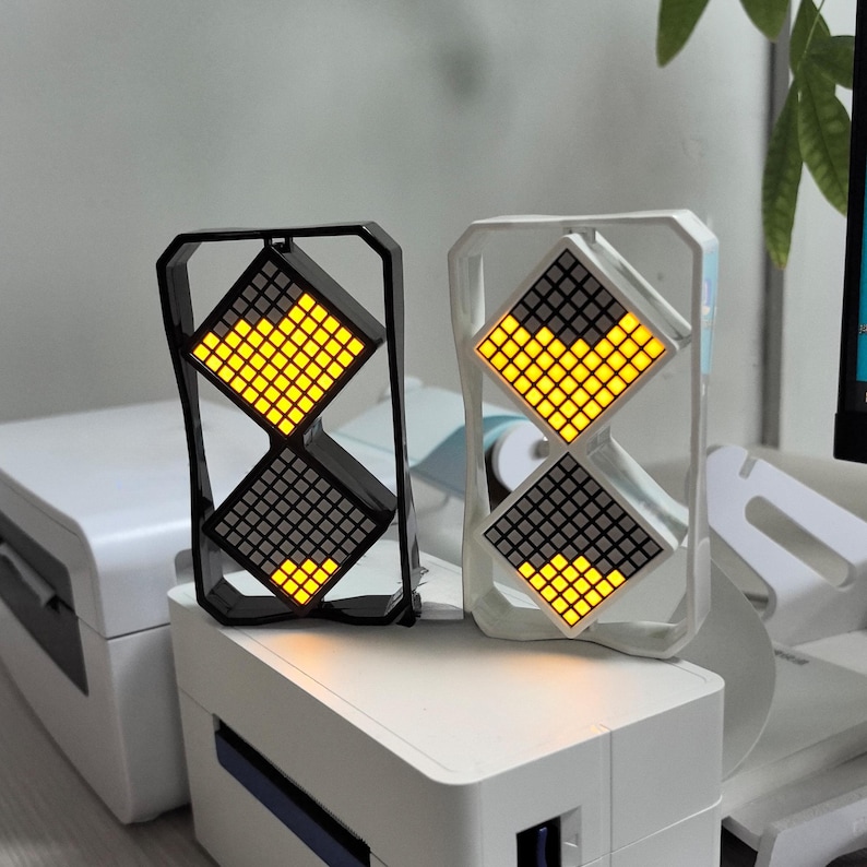 May include: Two digital clocks with a unique design. One clock has a black frame, the other is white. Each clock features two diamond-shaped displays with yellow and gray illuminated squares. The clocks are placed on a white surface.