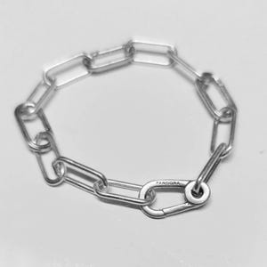 Könnte beinhalten: Ein silberfarbenes Kettenarmband aus länglichen, rechteckigen Gliedern. Das Armband verfügt über ein markantes, größeres Verschlussglied, das mit dem Wort PANDORA graviert ist und das runde Ende des Verschlusses mit den Kettengliedern verbindet.