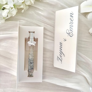 Può includere: Un flacone di profumo in vetro trasparente con dettagli argentati e un fiocco bianco, presentato in una scatola bianca. Un biglietto bianco con i nomi "Zeyna & Emren" in corsivo è accanto alla scatola. Lo sfondo presenta fiori e tessuto bianchi.