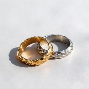 Könnte beinhalten: Zwei Metallringe mit einem strukturierten, geflochtenen Muster auf der Außenfläche. Ein Ring ist in einem polierten Goldton gehalten, während der andere Ring in einem polierten Silberton ausgeführt ist. Beide Bänder zeigen ein ineinandergreifendes Design.