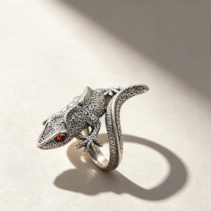 May include: A silver ring shaped like a lizard with a textured, scaly surface and a single small, round, deep red gemstone set as the eye. The lizard's body curves to form the band of the ring, with its tail extending upwards and outwards.
