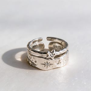 May include: A silver-colored open ring featuring a textured, hammered surface. The wider band displays a small, engraved star with a central clear stone, while the thinner upper band features a small star-shaped charm with a clear stone. S925 is engraved.