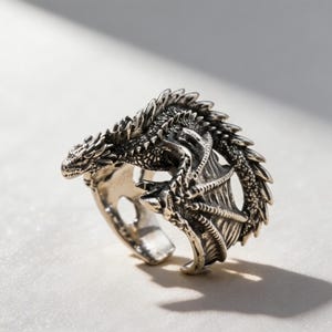 May include: A silver-toned metal ring featuring a detailed dragon design that wraps around the finger. The dragon has textured scales, prominent spikes along its back, and folded wings that form the band of the ring with an open back design.