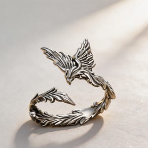 May include: A silver-toned metal ring shaped like a phoenix with wings spread and a long, curved tail. The design features intricate, raised feather detailing throughout the body and wings, with a polished finish that highlights the contours.