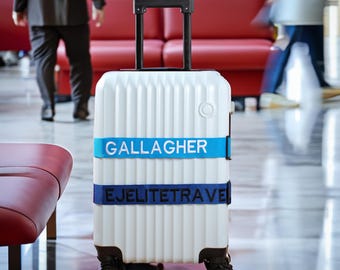 May include: A white rolling suitcase with vertical ribbed detailing and a retractable black handle. The suitcase has two blue bands with the text "GALLAGHER" and "EJELITETRAVE". The suitcase has four black wheels.