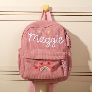 May include: Pink corduroy backpack with the name "Maggie" embroidered in white script. Features a front pocket with a rainbow and heart design, and a small tassel. The backpack is hanging from a yellow heart-shaped hook.