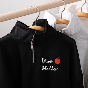 May include: A black quarter-zip sweatshirt featuring a silver metal zipper and an embroidered design on the chest. The embroidery includes the white text Mrs. Stella positioned above a small red apple with a green leaf, all on a solid black fabric.