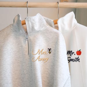 Pode incluir: Duas camisolas de quarto de fecho penduradas numa barra de madeira. Uma é cinzenta clara com "Mrs. Avery" bordado em letras douradas, um gráfico de abelha e um fecho de correr prateado. A outra é branca com "Mr. Smith" e um gráfico de maçã vermelha.