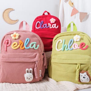 May include: Three corduroy backpacks in pink, red, and green, each with a name embroidered in colourful thread: Perla, Clara, and Chloe. The backpacks have a flower and animal-shaped embellishments.