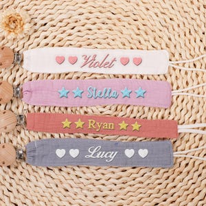 May include: Four personalized baby pacifier clips in white, pink, coral, and gray, each with a name and decorative accents. The clips have wooden beads and white cords, set on a woven background.