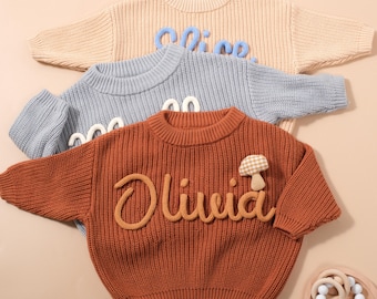 May include: Three knitted sweaters in beige, blue, and rust colors. The beige sweater has the name "Alice" in blue. The blue sweater has the name "Alice" in white. The rust sweater has the name "Olivia" in beige and a mushroom applique.