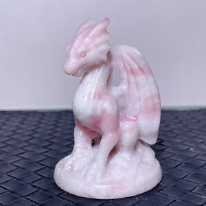 Pode incluir: Uma estatueta esculpida de um dragão em tons de rosa e branco. O dragão tem asas detalhadas e está empoleirado em uma base redonda. A escultura é feita de um material semelhante a pedra. As asas do dragão têm um padrão listrado.
