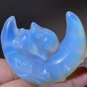 May include: A light blue, translucent sculpture of a sleeping cat nestled in a crescent moon. The cat and moon are carved with intricate details, and the sculpture has a smooth, polished finish. The moon has a few small, triangular shapes carved into it.