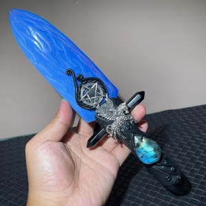 Puede incluir: Una daga de fantasía con una hoja azul y adornos plateados intrincados. El mango presenta una base negra con una piedra de labradorita y un diseño detallado. Un pentagrama es visible en la guarda.