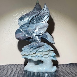 May include: A carved stone sculpture of an eagle with outstretched wings. The sculpture is primarily blue and gray, with detailed feather carvings. The eagle is perched atop a stylized tree, creating a decorative art piece.
