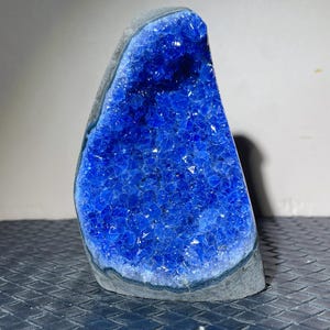 May include: A large, triangular geode with a vibrant, deep blue crystal interior. The outer edge is a dark gray, contrasting with the sparkling blue crystals. The geode is displayed upright, showcasing its natural form.