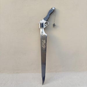 Puede incluir: Una espada plateada única con un mango estilo revólver. La hoja presenta un diseño grabado y una cadena decorativa con un dije. El mango es gris y plateado. El diseño general es una fusión de armas.