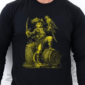 以下が含まれることがあります： 黒い長袖シャツ。黄色いイラストは、海賊猫とオウム。帽子をかぶった猫が樽の上に立ち、剣を持っています。ヴィンテージスタイルのイラストです。