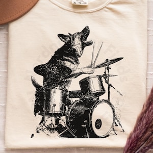 Könnte beinhalten: Cremefarbenes T-Shirt mit einer Schwarz-Weiß-Illustration eines Hundes, der ein Schlagzeug spielt. Der Hund ist mit offenem Maul dargestellt, als würde er singen. Das Shirt trägt den Text "COMFORT COLORS" und "XL - TG - EG".