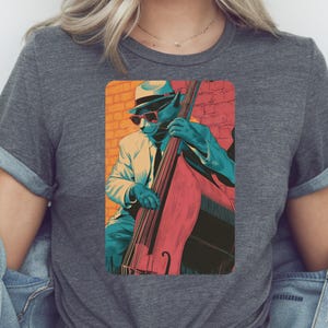 Puede incluir: Camiseta gris con una ilustración colorida de un gato tocando un contrabajo. El gato lleva traje, sombrero y gafas de sol, sobre un fondo de pared de ladrillos. Una prenda casual para el día a día.