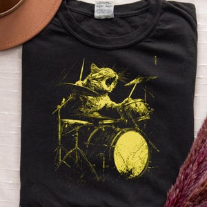 Może przedstawiać: Czarny t-shirt z żółtą ilustracją kota grającego na perkusji. Kot jest przedstawiony w trakcie ryczenia, z pałeczkami w łapach. Zestaw perkusyjny zawiera talerze i bęben basowy. Brązowy kapelusz i suszone kwiaty znajdują się w tle.