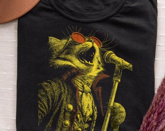 May include: Black t-shirt featuring a yellow illustration of a cat wearing sunglasses and a coat, singing into a microphone. The cat has a detailed design with orange sunglasses and a yellow microphone.