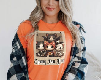 Pode incluir: Camiseta laranja com um gráfico de quatro gatinhos de desenho animado vestidos com fantasias de Halloween. A imagem inclui um morcego, uma múmia e um gatinho com uma fantasia de abóbora. O texto "Spooky Paw Kittys" está abaixo.
