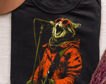 May include: Black t-shirt featuring a graphic of a cat singing into a microphone. The cat wears red sunglasses and an orange jacket. The design has a vintage, distressed look. The shirt is a crew neck and appears to be made of cotton.