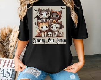 Pode incluir: Camiseta preta com um design de Halloween. O design mostra quatro gatos de desenho animado fantasiados, incluindo uma múmia e um vampiro, com morcegos e o texto "Spooky Paw Kittys". Os gatos estão empilhados.