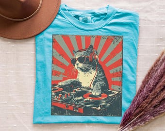 Puede incluir: Una camiseta turquesa con un gráfico de un gato con gafas de sol y auriculares, operando una mesa de mezclas de DJ. El diseño tiene una estética retro y desgastada con acentos rojos y grises.