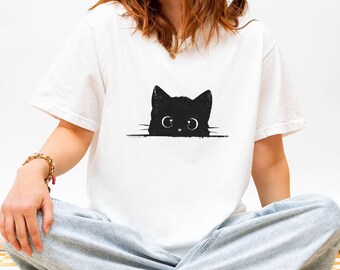 May include: White t-shirt featuring a black cat graphic. The cat's head is visible above a horizontal black line, with large eyes and a small pink nose. The shirt is a casual style.
