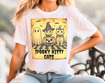 Może przedstawiać: Biała koszulka z motywem Halloween. Projekt przedstawia ducha, dwa koty i dynię. Tekst "Spooky Kitty Cats" jest wydrukowany poniżej. Koty noszą kapelusz czarownicy i muszkę.
